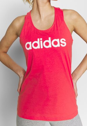 Kobieta w jaskrawoczerwonym, bezrękawniku Adidas z białym logo, ręce na biodrach, ubrana w jasno szare spodnie, zdjęcie przycięte na wysokości szyi i ud.