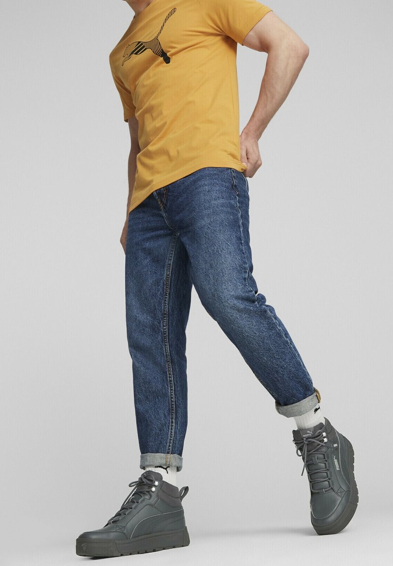 Homme portant un t-shirt jaune, un jean bleu retroussé, des chaussettes blanches et des baskets montantes gris foncé, debout avec une jambe légèrement levée.