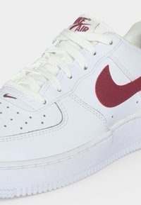 Λευκό αθλητικό παπούτσι Nike Air με σκούρο κόκκινο σήμα, λευκά κορδόνια, διατρημένο μπροστινό μέρος και λευκή σόλα, εμφανίζεται σε κοντινό πλάνο.