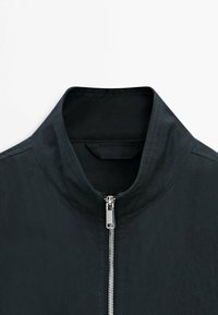 Giacca nera con colletto alla coreana e cerniera frontale argento, esposta su uno sfondo bianco semplice.