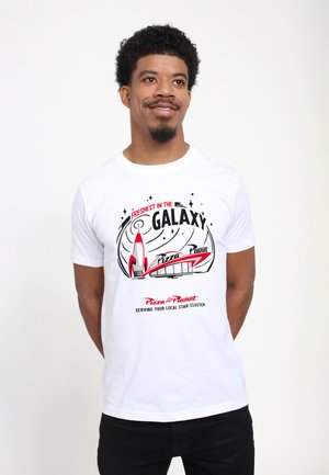 Homme aux cheveux courts et bouclés avec une barbiche, portant un T-shirt blanc avec un graphisme rouge et noir "Freshest in the Galaxy Pizza Planet", souriant et debout.