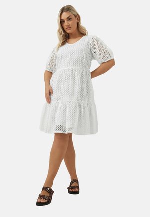 Vestido blanco de longitud media con un diseño de tejido abierto texturizado, mangas cortas abullonadas y un dobladillo acampanado. Combinado con sandalias marrones con tachas.
