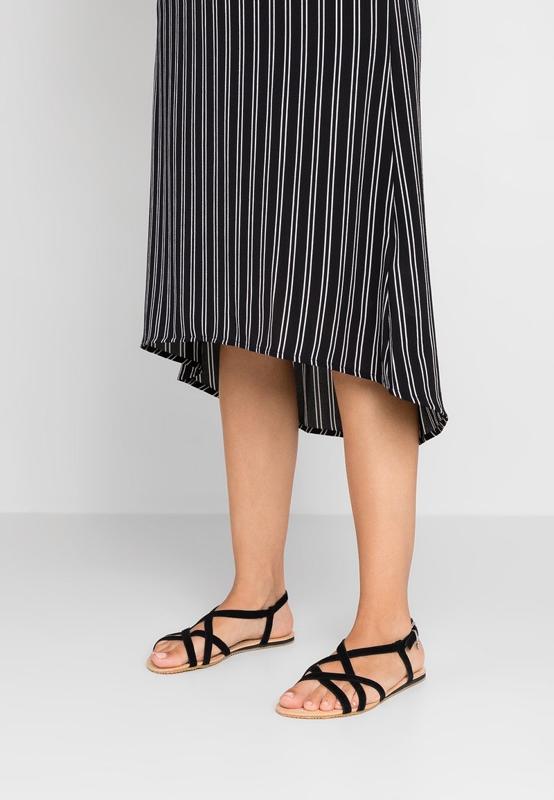 Personne portant des sandales noires à multiples brides et une jupe midi rayée verticalement noire et blanche avec un ourlet asymétrique.