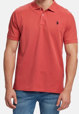 Homme portant un polo rouge à manches courtes avec deux boutons et un jean bleu, debout devant un fond blanc uni.