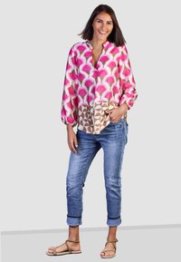 Bunte Bluse mit rosa und braunen geometrischen Mustern, kombiniert mit hochgekrempelten Jeans und braunen Riemchensandalen.
