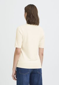 Femme aux cheveux bruns mi-longs portant un haut crème à manches courtes et un jean bleu foncé, debout devant un fond clair uni.