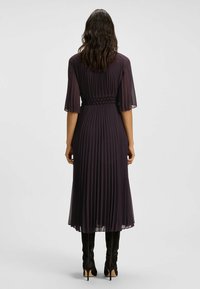 Violetti laskostettu mekko, jossa on lyhyet liehuttavat hihat ja smokattu vyötärö, yhdistetty mustiin polven korkeisiin korkokenkiin, esitettynä takaa.