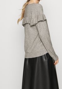 Femme portant un pull en tricot gris clair avec un détail en volants dans le dos et une jupe en cuir noir, se tenant devant un fond uni.