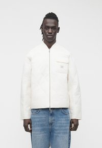 Balta dygsniuota striukė su užtrauktuku priekyje, turi krūtinės kišenę ir plačias rankoves, derinama su šviesiai mėlynais džinsais. Lygi tekstūra.