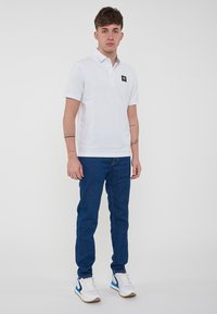Polo shirt bianco con un piccolo logo nero, abbinato a jeans blu e sneakers bianche con accenti blu. Design pulito, look aderente.