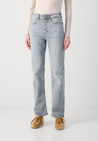 Jeans azzurri chiari a vita alta con taglio dritto, dotati di chiusura con bottone e cinque tasche, abbinati a scarpe marrone chiaro.