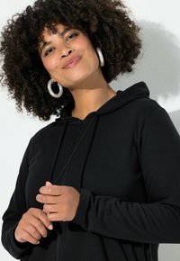 Mujer con cabello rizado que lleva pendientes de aro de plata y una sudadera con cremallera negra sonriendo suavemente contra un fondo blanco.