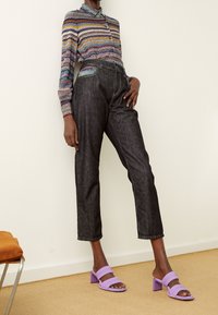 Person som bär mörka korta jeans, en långärmad topp med flerfärgad zigzag-mönstring och lavendelblock-klacksandaler som står inomhus.