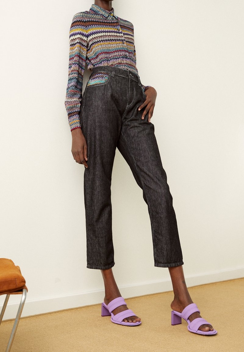 Person som bär mörka korta jeans, en långärmad topp med flerfärgad zigzag-mönstring och lavendelblock-klacksandaler som står inomhus.