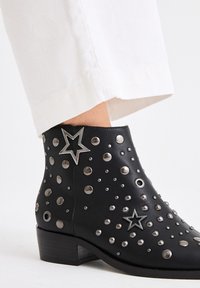 Botas de tobillo de cuero negro adornadas con tachuelas plateadas y un acento de estrella. Presentan un tacón bajo y una textura suave.