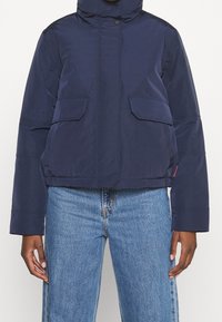 Person som bär en marinblå vadderad jacka med framfickor och vida blå jeans mot en enhetlig bakgrund.