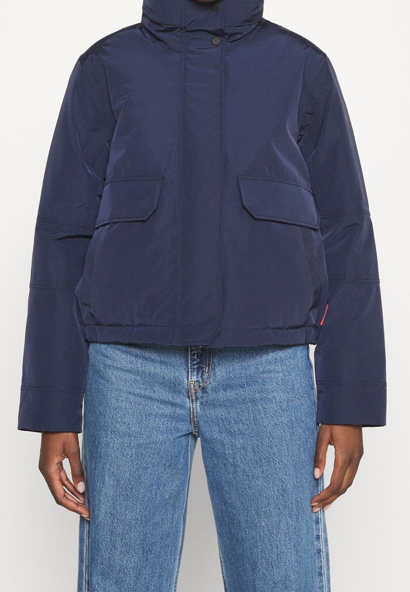 Person som bär en marinblå vadderad jacka med framfickor och vida blå jeans mot en enhetlig bakgrund.