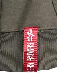 Olivgrüne Kapuzenjacke mit geripptem Muster; verfügt über ein auffälliges rotes Etikett mit weißem Text und einem Logodesign am Saum.
