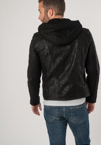 Giacca nera in pelle sintetica con cappuccio, caratterizzata da una superficie texturizzata e dettagli delle cuciture, abbinata a jeans blu.