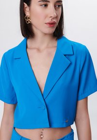 Camisa azul corta con cuello en V profundo, mangas cortas y una textura suave. Presenta una pequeña etiqueta con el logo en el dobladillo.