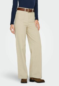 Pantalons larges beiges en tissu texturé, avec des poches avant, une ceinture marron avec une boucle dorée et des bottines marron.