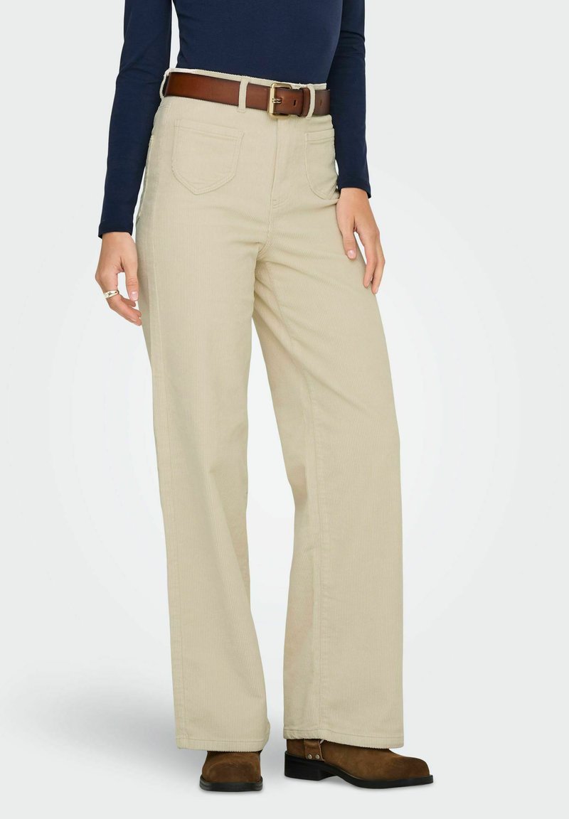 Pantalons larges beiges en tissu texturé, avec des poches avant, une ceinture marron avec une boucle dorée et des bottines marron.