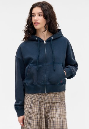 Sudadera de cremallera azul marino hecha de tela suave. Cuenta con un bolsillo frontal, cordones ajustables y un corte holgado con puños de canalé.