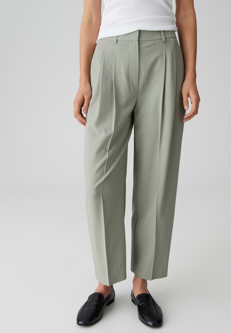 Persoon draagt lichtgroene broek met rechte pijpen, zwarte loafers en een witte mouwloze top tegen een effen lichte achtergrond.