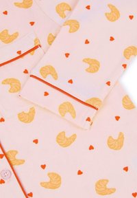 Pyjama-top van zacht roze stof met een patroon van gele croissants en rode harten. Oranje afwerking langs de kraag en manchetten.