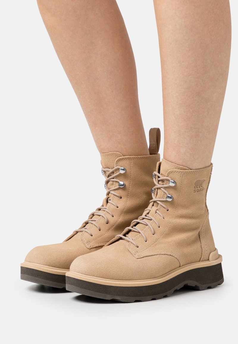 Beige Wildleder-Ankle-Boots mit Schnürung und silbernen Ösen, mit einer strukturierten Oberfläche und einer stabilen dunklen Sohle mit leichtem Plateau.