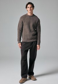 Pulover tricotat maro cu guler rotund, asociat cu pantaloni negri lejeri și pantofi maro deschis, creând un aspect relaxat.