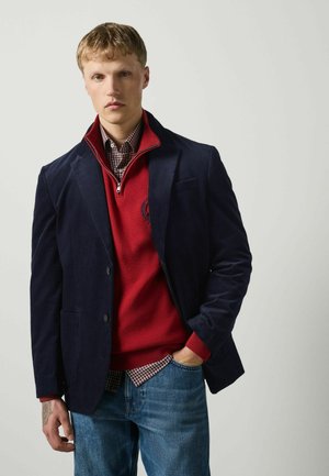 Blond man wearing a navy blazer over a red quarter-zip sweater and checkered shirt, with blue jeans and one hand in pocket.