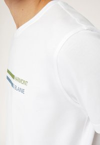 T-shirt bianco in cotone con maniche corte. Il testo in verde e blu recita “HARMONT & BLAINE,” con righe orizzontali sotto il testo.
