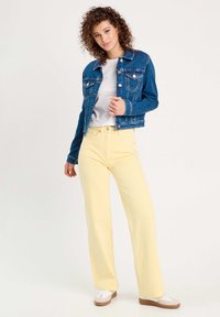 Chaqueta de mezclilla en azul oscuro con botones plateados y bolsillos, combinada con pantalones amarillos de pierna ancha de talle alto y zapatos blancos con suelas beige.