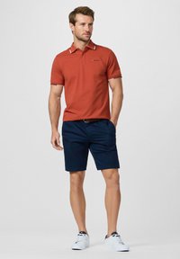 Homme debout, la main gauche dans la poche de son short bleu marine, portant un polo couleur rouille et des baskets blanches sur un fond uni.