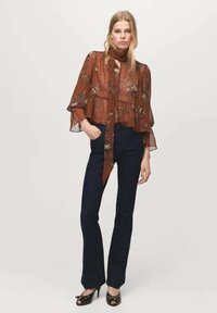 Zwiewna, przezroczysta brązowa bluzka w kwiaty z rozkloszowanymi rękawami i wiązaniem przy dekolcie, zestawiona z ciemnoniebieskimi dopasowanymi dżinsami i czarnymi butami z kokardkami.