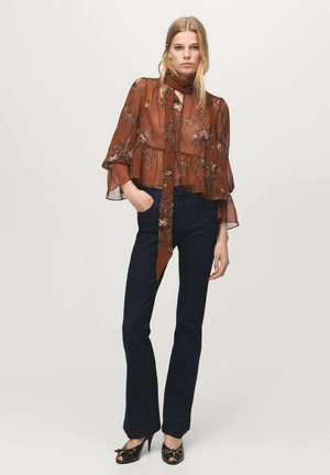 Blusa marrón floral, ligera y transparente, con mangas acampanadas y un lazo en el cuello, combinada con jeans ajustados azul oscuro y zapatos negros con lazos.