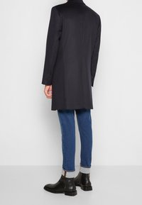 Cappotto in misto lana navy con maniche lunghe, vestibilità sartoriale, rever a punta e polsini con bottone, abbinato a jeans blu e stivali alla caviglia in pelle nera.