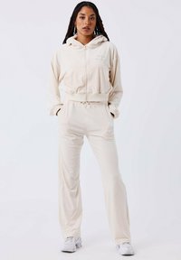 Beige zip-up hoodie met een kangoeroezak en bijpassende broek, gemaakt van zachte stof. De capuchon heeft een trekkoord en is ontworpen voor comfort.