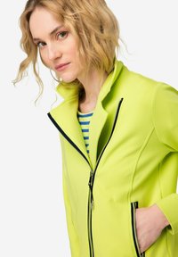 Limoengroene ritsjacket van gladde stof, met een hoge kraag, zwarte ritsen en zijzakken. Gestreept shirt eronder.