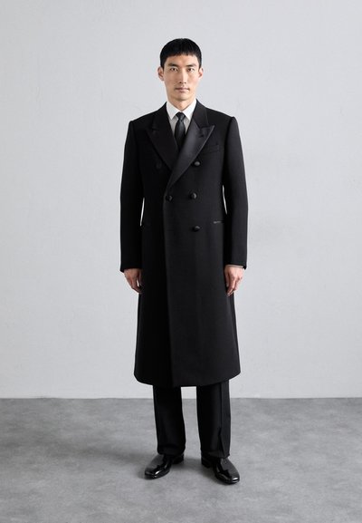 Abrigo negro de doble botonadura con solapas de satén, ajuste entallado, hombros estructurados y textura suave. Llevado sobre pantalones y zapatos a juego.