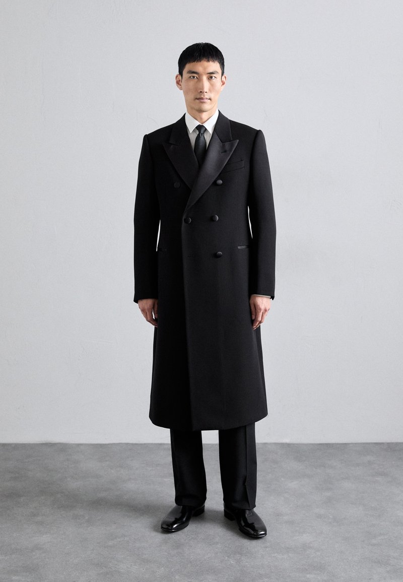 Melnā divrindu mētelis ar satīna atlokiem, pielāgota silueta, stingri pleci un gluda tekstūra. Valkāts pār atbilstošām biksēm un apaviem.