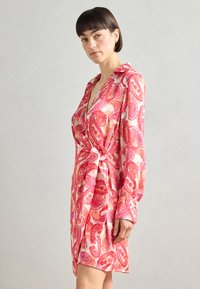 Robe portefeuille à motifs paisley roses, avec des manches longues, une ceinture à nouer et un col. Tissu doux avec une texture soyeuse. Longueur courte.