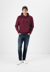 Burgunderfarget hettegenser med frontlomme og hvit logo, kombinert med blå slim-fit jeans og hvite sneakers. Modellen har på seg en mønstret caps.