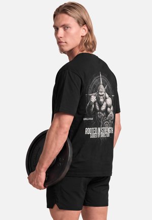 Jonge man met schouderlang haar houdt een gewichtplaat vast, draagt een zwart T-shirt met een gespierde gorilla-afbeelding en tekst op de achterkant.