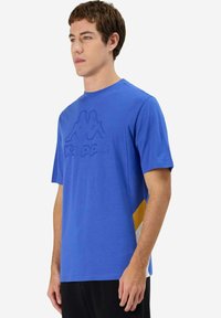 Camiseta de manga corta azul con un logo texturizado. Presenta un panel lateral con acentos en amarillo y blanco. Fabricada con una tela suave y transpirable.
