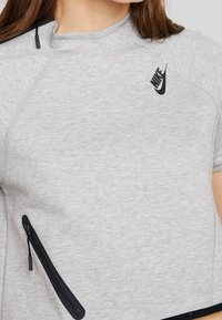 Šedé sportovní tričko s krátkým rukávem, kulatým výstřihem, černým logem Nike a černou kapsou na zip na boku. Měkká textura, ležérní design.