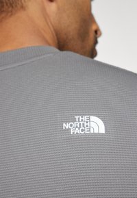 Siva teksturna tkanina košulja s bijelim logom "THE NORTH FACE" na gornjem dijelu leđa, s dizajnom okruglog izreza.