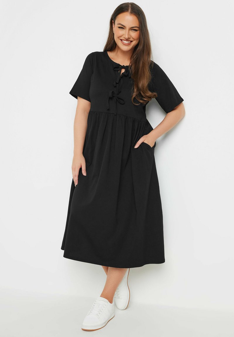 Mustahap musta kleit lühikeste varrukatega, vöökohaga ja paeltega kaela juures, ning küljetaskud; kantud valgete tuhvlitega.