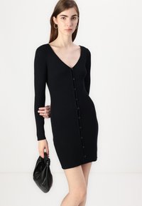 Robe noire côtelée avec un décolleté en V, des manches longues et des détails de boutons sur le devant. Modèle tenant un sac à main en tissu noir.